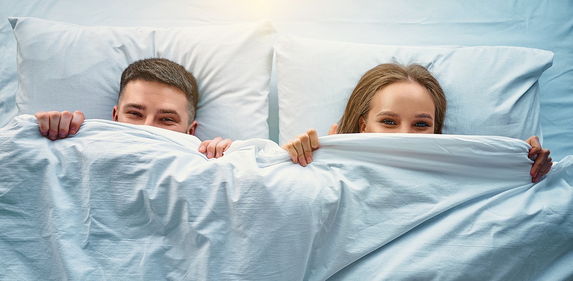 Junges lächelndes Paar liegt nebeneinander im Bett. Beide ziehen sich die weiße Bettdecke halb über das Gesicht