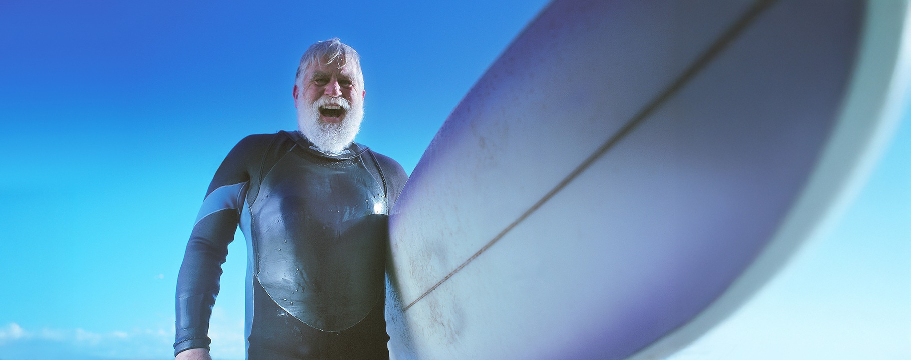 Älterer Herr mit weißen Haaren und weißem Vollbart im Neoprenanzug, ein Surfbrett unter dem Arm tragend, bei strahlend blauem Himmel am Meer
