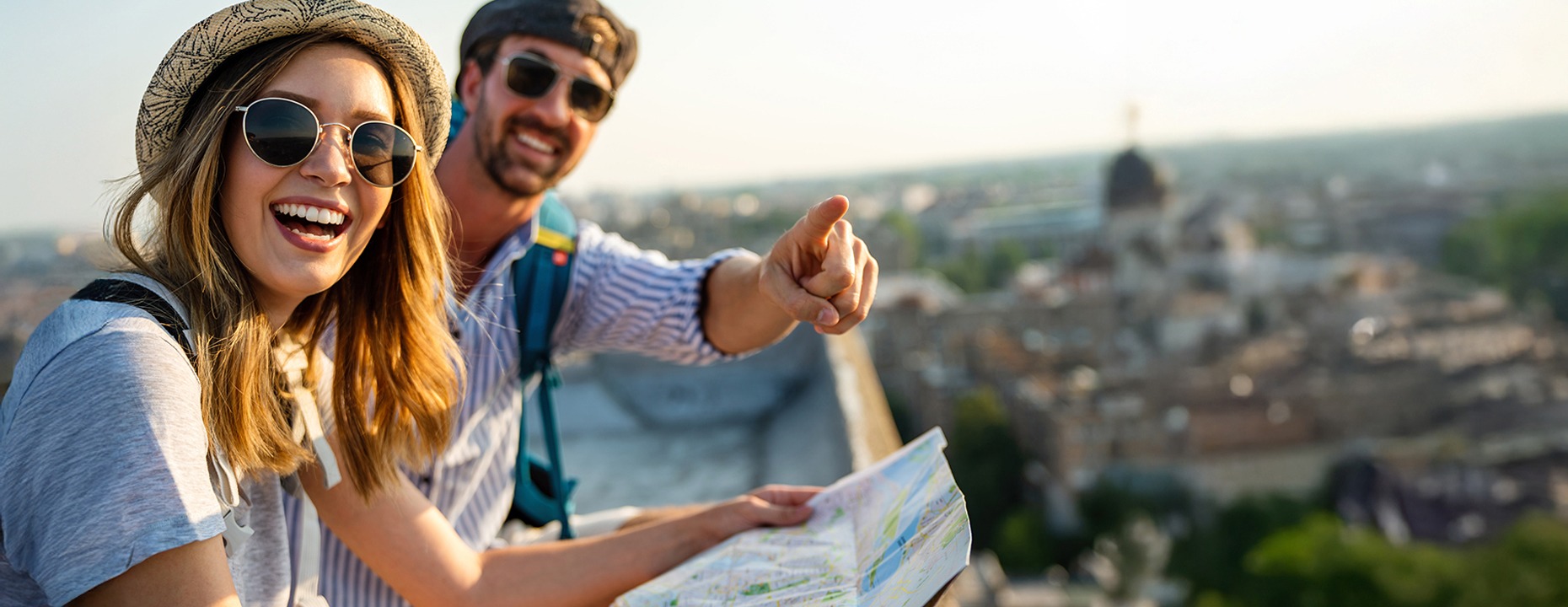 Junges Touristen-Paar mit Sonnenbrillen, Kopfbedeckungen und Rucksäcken auf einer Aussichtsplattform über einer Stadt bei Sonnenschein.