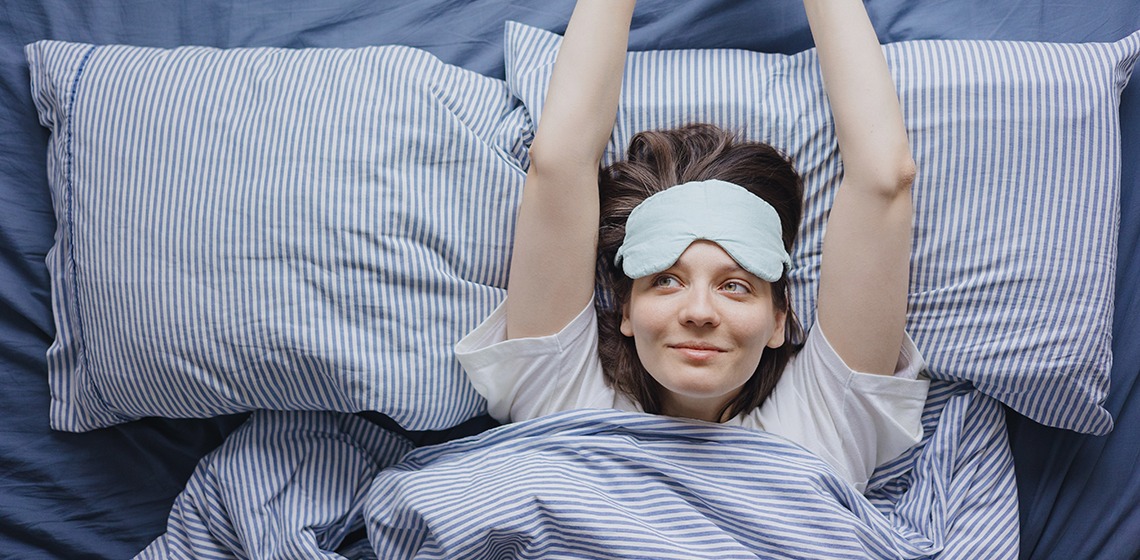 Eine Frau die sich im Bett streckt und eine Schlafmaske an hat