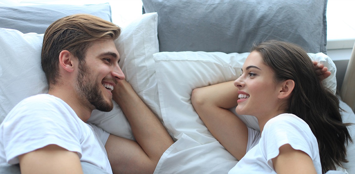 Junger Mann und junge Frau liegen sich im Bett in Seitenlage gegenüber und lächeln sich zufrieden an